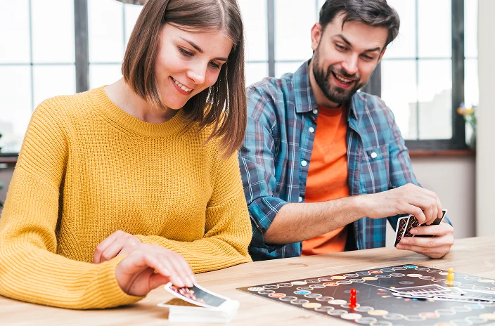 Een glimlachende vrouw en man spelen een bordspel, zittend aan een houten tafel. Zij draagt een gele trui; hij heeft een geruit overhemd aan. Ze zien er vrolijk en betrokken uit.